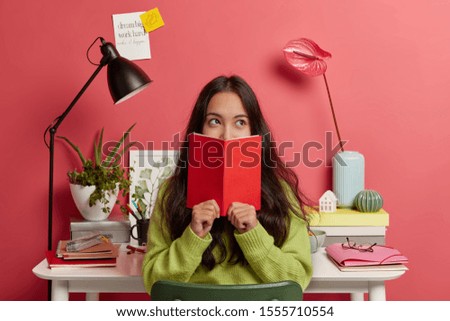 Beautiful brunette pensive mixed race female student learns information from textbook, covers half of face with red diary, poses in coworking space against desktop with necessary things for studying