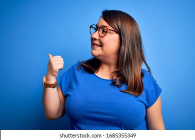 Schöne Brunette plus Größe Frau mit lockerem T-Shirt auf isoliertem blauen Hintergrund lächeln mit glücklichem Gesicht und zeigen auf die Seite mit Daumen nach oben.