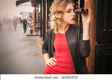 Beautiful Brunette Sexy Spy Agent (killer Or Police) Woman In Leather Jacket And Red Luxury Dress With A Binocular In Her Hand Stalking Someone, To Catch Him In European City.