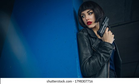 Beautiful brunette sexy spy agent (killer or police) woman in leather jacket and jeans with a gun in her hand running after someone, to catch him on parking.