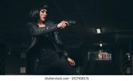 Beautiful brunette sexy spy agent (killer or police) woman in leather jacket and jeans with a gun in her hand running after someone, to catch him on parking.