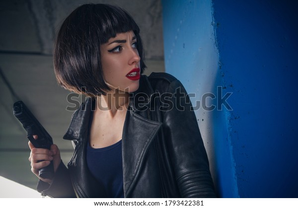 Beautiful brunette sexy spy agent (killer\
or police) woman in leather jacket and jeans with a gun in her hand\
running after someone, to catch him on\
parking.