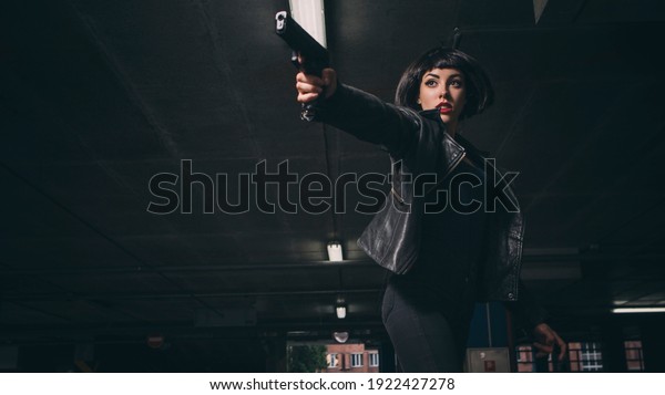 Beautiful brunette sexy spy agent (killer\
or police) woman in leather jacket and jeans with a gun in her hand\
running after someone, to catch him on\
parking.