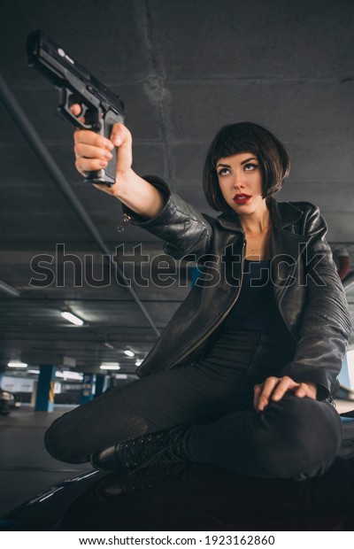 Beautiful brunette sexy spy agent (killer
or police) woman in leather jacket and jeans with a gun in her hand
running after someone, to catch him on
parking.