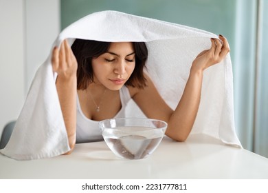 Beautiful brunette short-haired young lady doing facial streaming procedure at home, sit at vanity table above bowl with hot water and essential oils, cover head with bath towel. Domestic skin care