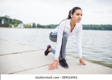 Beautiful Brunette Sporty Caucasian Female Woman Start To Running Outdoors. Fit Woman Doing Marathon Training. Fitness Motivation. Healthy Athlete Girl With Earphones Stretching In Park Near To Lake.