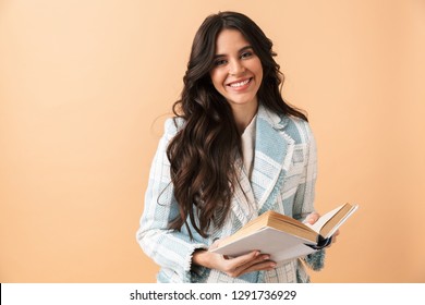 Beautiful brunette woman dressed in plaid jacket standing isolated over beige background, holding a book