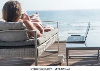 Beautiful Brunette Woman Freelancer Wrapped In A Plaid Resting On A Couch And Sunbathing On The Terrace With A Cup Of Coffee At Summer Day. Sea View. WFH. Working From Home 