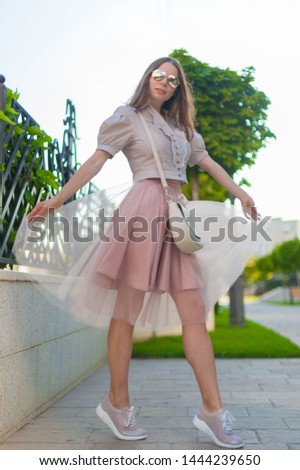 Beautiful brunette woman, has peach color net skirt, beige blouse, sunglasses, leather handbag, sneakers . Urban city portrait. People in street. Happy concept. Summer fashion style. 