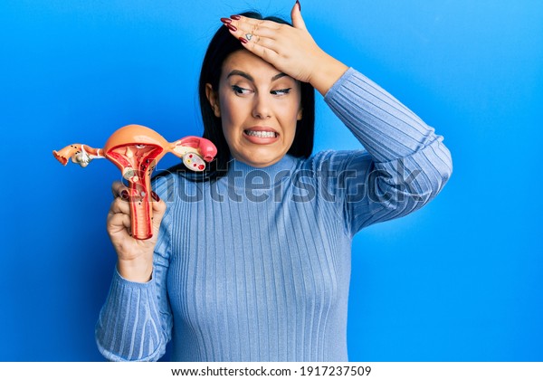 Beautiful brunette woman holding anatomical model of\
female genital organ stressed and frustrated with hand on head,\
surprised and angry face\
