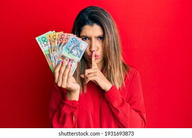 Beautiful brunette woman holding australian dollars asking to be quiet with finger on lips. silence and secret concept. 