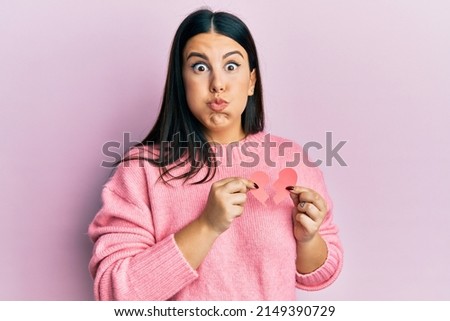Beautiful brunette woman holding broken heart paper shape puffing cheeks with funny face. mouth inflated with air, catching air. 