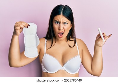 Beautiful Brunette Woman Holding Sanitary Pad And Cotton Tampon In Shock Face, Looking Skeptical And Sarcastic, Surprised With Open Mouth 