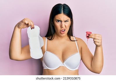 Beautiful Brunette Woman Holding Sanitary Pad And Reusable Menstruation Cup Clueless And Confused Expression. Doubt Concept. 