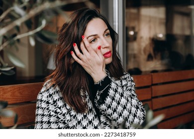 A beautiful brunette woman in a hounds tooth check coat sits in an outdoor cafe and thinking - Powered by Shutterstock