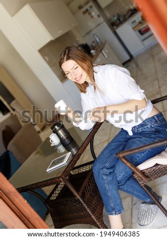 Beautiful brunette woman housewife in white t-shirt and jeans at home in the interior in the kitchen holding a cup of coffee in her hands