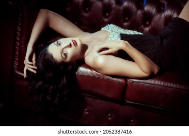 Beautiful brunette woman lying on a red sofa in an elegant evening dress with bright makeup and flowing wavy hair in a vintage interior background. Elegant evening style of the 1930s-1940s. 