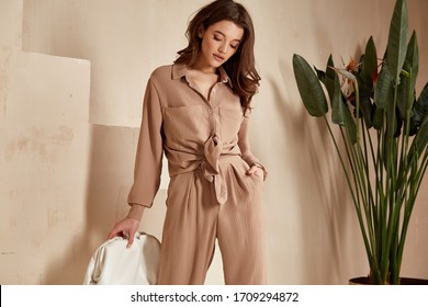 Beautiful brunette woman natural makeup wear fashion clothes casual dress code office style total beige blouse and pants suit, romantic date business meeting accessory bag interior stairs flowerpot.