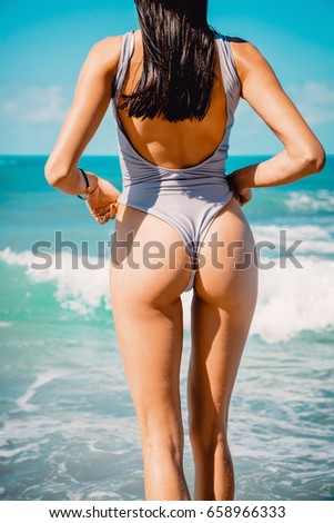 Beautiful brunette woman with perfect body standing up on beach, wearing stylish bikini and blue ocean