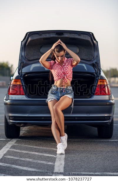 A beautiful brunette woman in shorts\
and a red shirt leaned on the trunk of her\
car.
