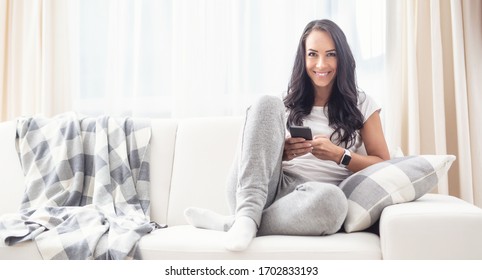Beautiful brunette woman sitting on a white sofa next to grey and white blanket and pillow, smiling to the camera, holding a phone.