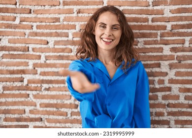 Beautiful brunette woman standing over bricks wall smiling friendly offering handshake as greeting and welcoming. successful business.  - Powered by Shutterstock