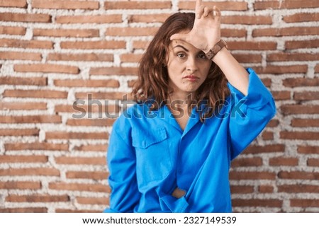 Beautiful brunette woman standing over bricks wall making fun of people with fingers on forehead doing loser gesture mocking and insulting. 