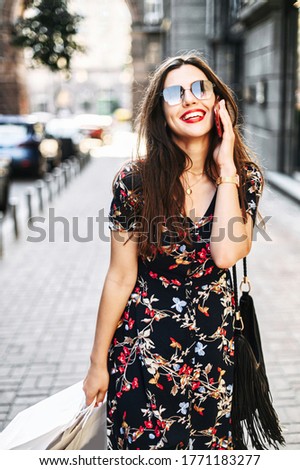 Beautiful brunette woman in sunglasses is talking on the phone outdoors on the city street, she is happy with the shopping and comes with paper bags in her hands. Vertical shot