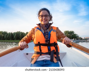 Beautiful brunette woman teen with glasses and in an orange life jacket rowing oars while sitting in a boat. Family walks at the boat station. Illuminated by sun glare - Powered by Shutterstock