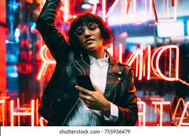 Beautiful brunette woman in trendy apparel and eyewear enjoying nightlife in city listening music in earphones and moving to sound, gorgeous hipster girl dancing outdoors on neon city illumination 