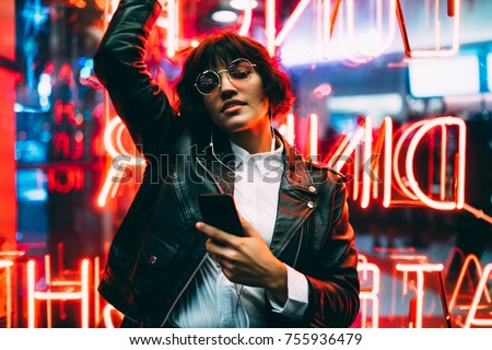 Beautiful brunette woman in trendy apparel and eyewear enjoying nightlife in city listening music in earphones and moving to sound, gorgeous hipster girl dancing outdoors on neon city illumination 
