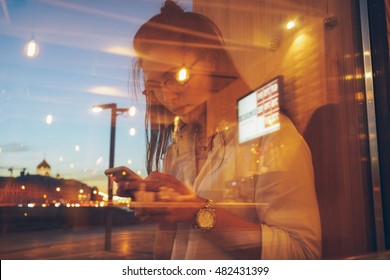 A Beautiful Brunette Woman Wearing Glasses And Looking At The Screen Of A Mobile Phone While Standing Behind The Student Cafe Window With A  Reflection Of The Sunset Colors. Blurred Photo.