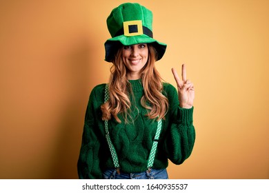Beautiful brunette woman wearing green hat with clover celebrating saint patricks day smiling with happy face winking at the camera doing victory sign. Number two.