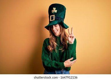 Beautiful brunette woman wearing green hat with clover celebrating saint patricks day smiling with happy face winking at the camera doing victory sign. Number two.