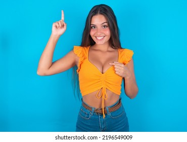 Beautiful Brunette Woman Wearing Orange Tank Top Over Blue Background Says: Wow How Exciting It Is, Has Amazed Expression, Indicates Something. One Hand On Her Chest And Pointing With Other Hand.