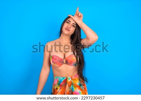 beautiful brunette woman wearing swimwear over blue background making fun of people with fingers on forehead doing loser gesture mocking and insulting.
