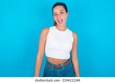 Beautiful Brunette Woman Wearing Withe Tank Top Shirt Over Blue Background Winking Looking At The Camera With Sexy Expression, Cheerful And Happy Face.