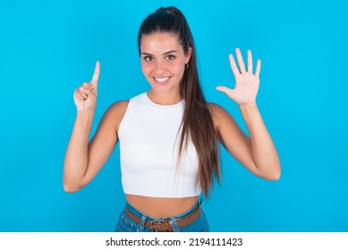 beautiful brunette woman wearing withe tank top shirt over blue background showing and pointing up with fingers number six while smiling confident and happy.