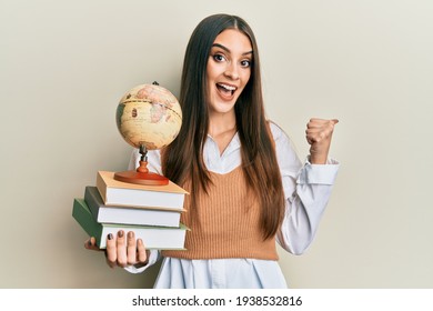 Beautiful brunette young woman holding pile of books and vintage world ball pointing thumb up to the side smiling happy with open mouth 