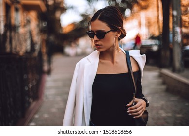 Beautiful Brunette  Young Woman With Low Bun Wearing Sunglasses, Circle Earrings, Black Top, White Blazer Or Jacket And Handbag Walking On The Street. Fashion Look. Sexy Business Woman. Sensual Girl