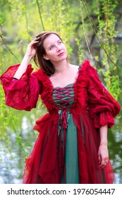 beautiful brunette young woman in red gown standing by lake under tree