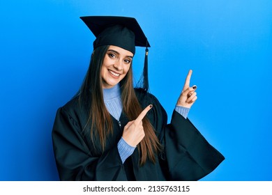 Beautiful brunette young woman wearing graduation cap and ceremony robe smiling and looking at the camera pointing with two hands and fingers to the side. 
