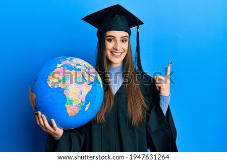 Beautiful brunette young woman wearing graduation robe holding world ball smiling happy pointing with hand and finger to the side 
