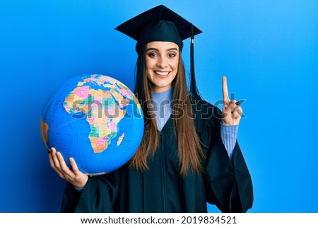 Beautiful brunette young woman wearing graduation robe holding world ball smiling with an idea or question pointing finger with happy face, number one 