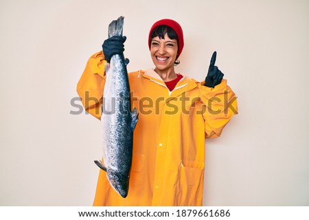 Beautiful brunettte fisher woman wearing raincoat holding fresh salmon surprised with an idea or question pointing finger with happy face, number one 