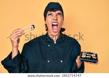 Beautiful brunettte woman chef holding sushi using chopsticks angry and mad screaming frustrated and furious, shouting with anger looking up. 