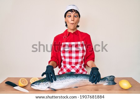 Beautiful brunettte woman fishmonger cooking fresh raw salmon making fish face with mouth and squinting eyes, crazy and comical. 