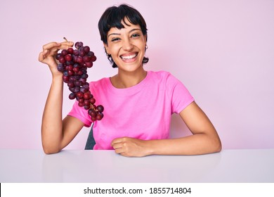 Beautiful brunettte woman holding branch of fresh grapes looking positive and happy standing and smiling with a confident smile showing teeth 