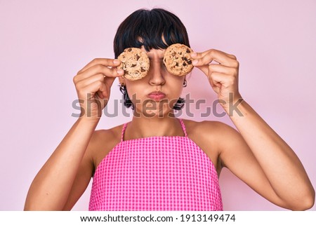 Beautiful brunettte woman holding chocolate chips cookies on eyes puffing cheeks with funny face. mouth inflated with air, catching air. 