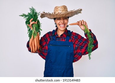 Beautiful brunettte woman wearing farmer clothes holding fresh carrots smiling and laughing hard out loud because funny crazy joke. 